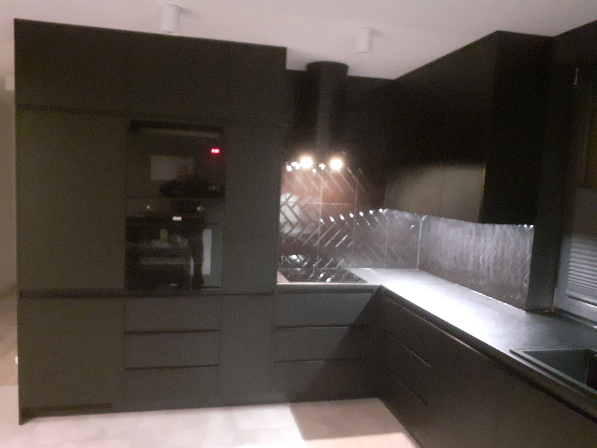 Nowoczesna, czarna kuchnia z matowymi frontami, piekarnikiem w zabudowie i płytkami chevron nad blatem. Minimalistyczny design z oświetleniem punktowym.