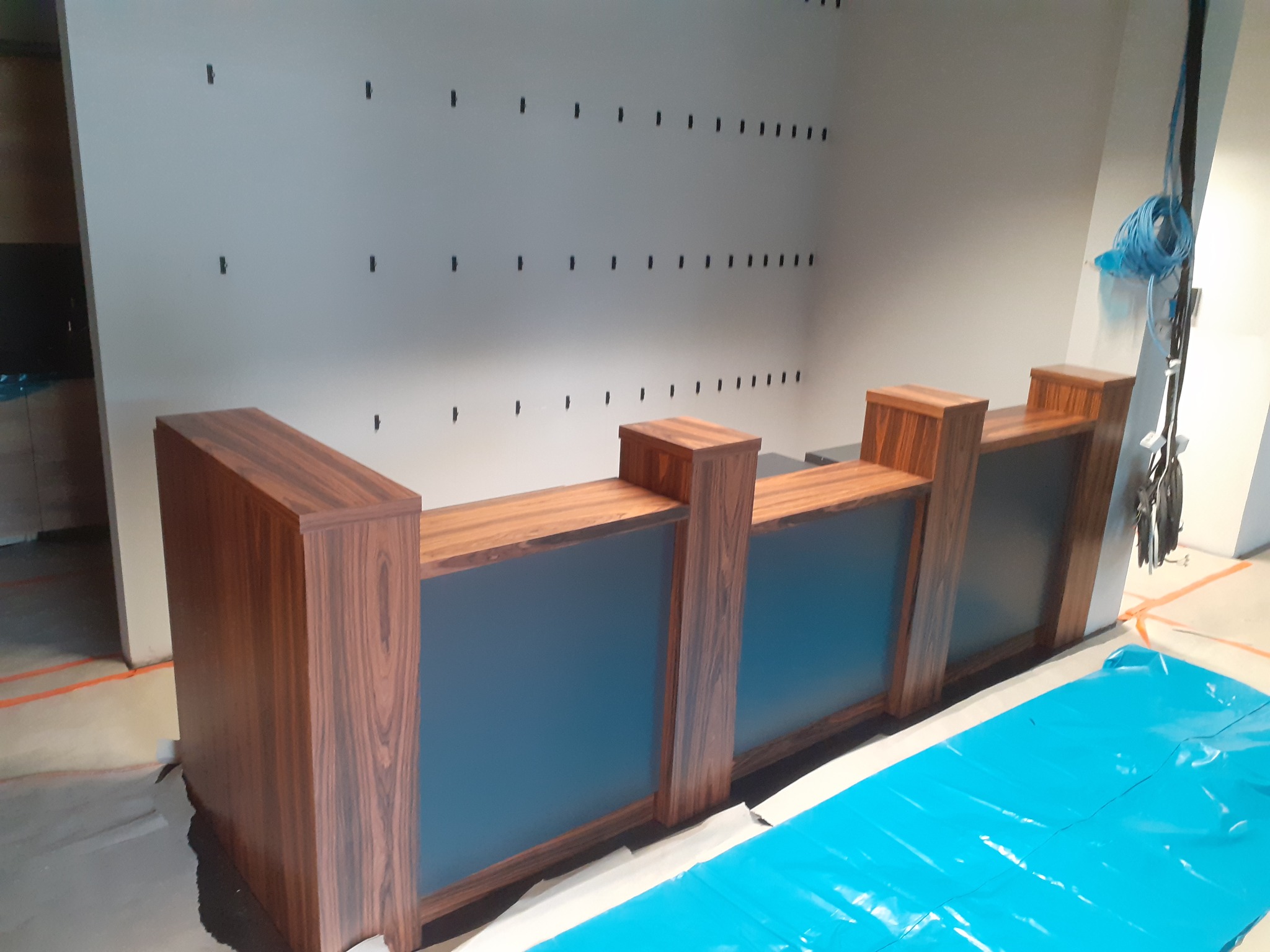 Nowoczesna lada recepcyjna z ciemnego drewna i niebieskiego szkła, zabezpieczona folią ochronną na podłodze, w surowym wnętrzu z widocznymi śladami po instalacjach.