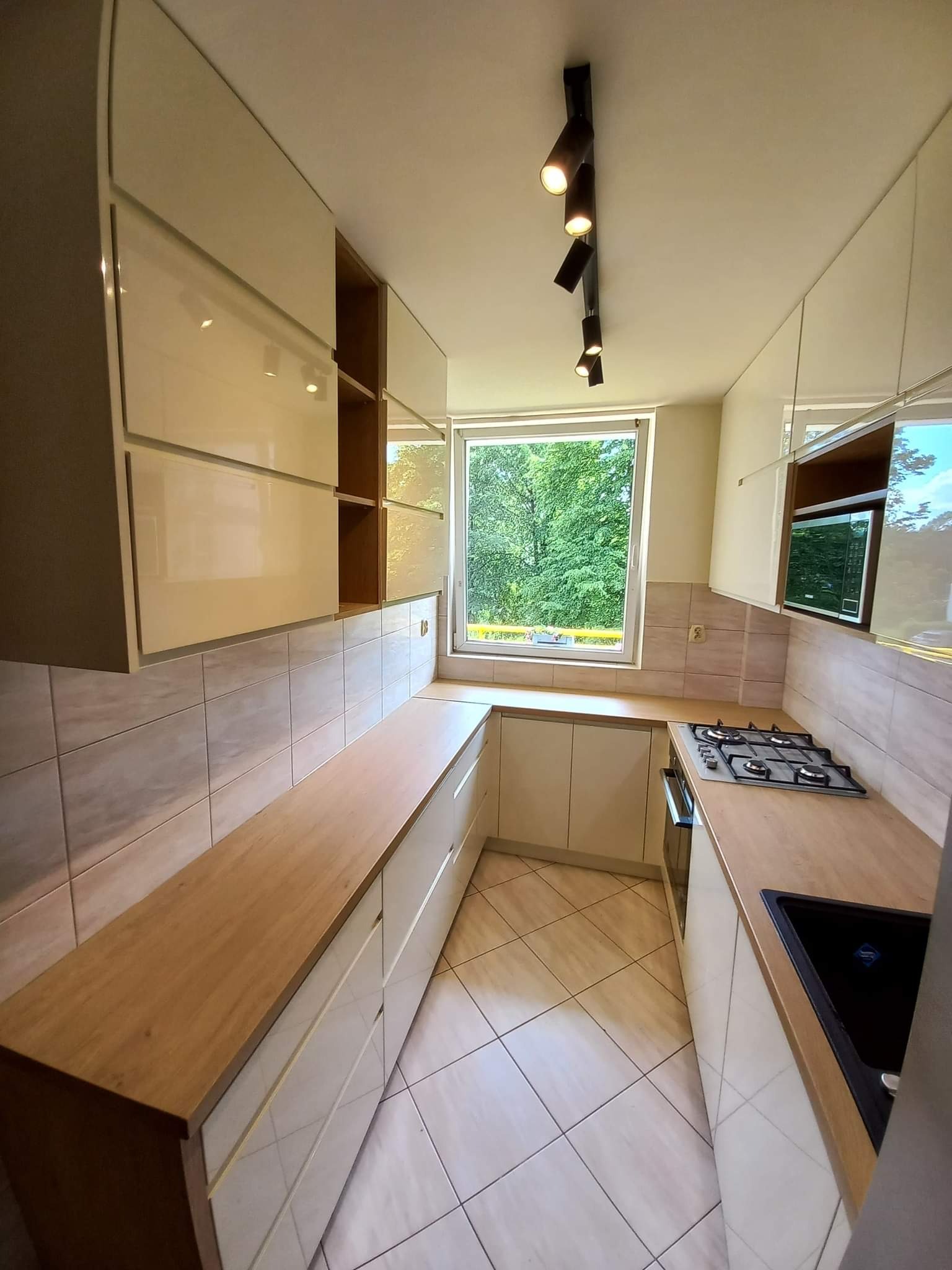 Jasna kuchnia z drewnianym blatem i widokiem na zieleń za oknem. Górne szafki lakierowane, oświetlenie punktowe na suficie. Ujęcie z perspektywy podłogi.