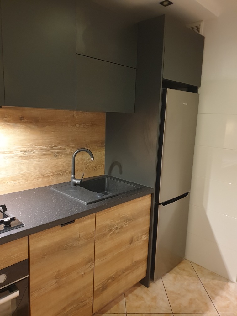 Nowoczesna kuchnia z szarymi szafkami górnymi i drewnopodobnymi dolnymi, czarnym zlewem i baterią, oraz srebrną lodówką Amica. Widok na fragment blatu i płytę grzewczą.