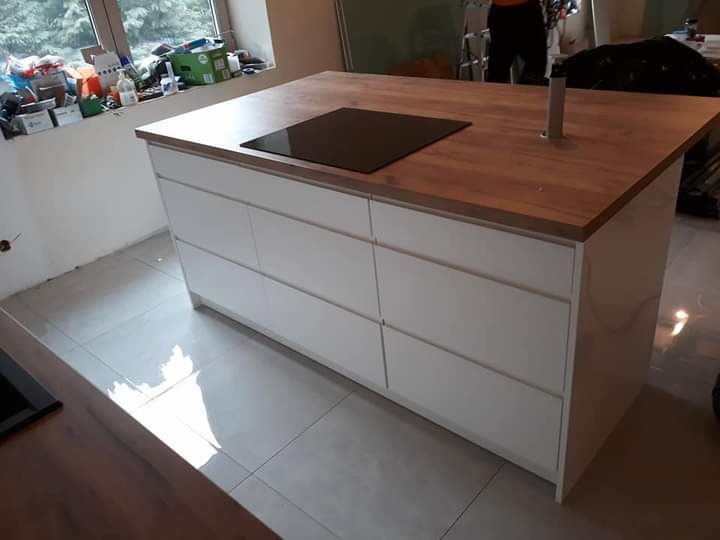 Wyspa kuchenna z drewnianym blatem i białą zabudową szuflad, z wbudowaną płytą grzewczą i wysuwanym gniazdkiem, na tle okna z akcesoriami i fragmentu pomieszczenia w trakcie wykończenia.