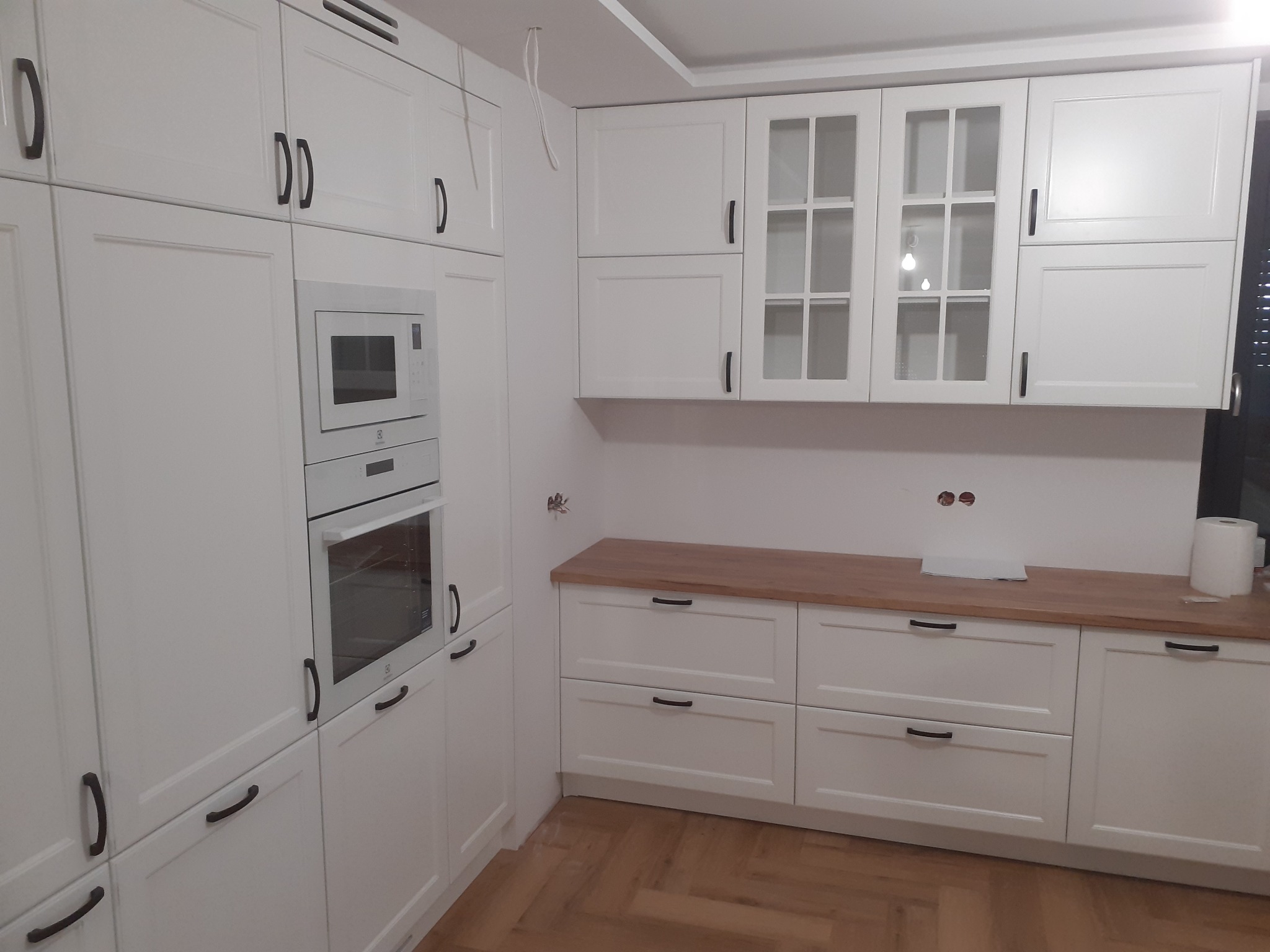 Biała kuchnia z drewnianym blatem, czarnymi uchwytami i wbudowanym sprzętem AGD. Widoczne szafki z przeszkleniami i oświetlenie sufitowe. Podłoga z paneli.