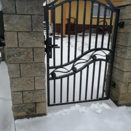 JSMP SYSTEM SPÓŁKA Z OGRANICZONĄ ODPOWIEDZIALNOŚCIĄ - Otwarta, czarna, metalowa furtka wejściowa z dekoracyjnymi elementami, osadzona pomiędzy dwoma kamiennymi słupkami, pokryta śniegiem w zimowej scenerii.