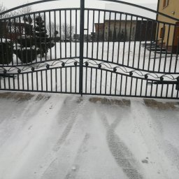 JSMP SYSTEM SPÓŁKA Z OGRANICZONĄ ODPOWIEDZIALNOŚCIĄ - Czarna metalowa brama wjazdowa z dekoracyjnymi elementami w kształcie kulek, widok z zewnątrz na ośnieżony podjazd, w tle budynek z czerwonej cegły i zielone drzewa.