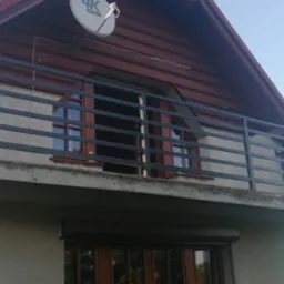 Szara, metalowa balustrada balkonowa z poziomymi szczeblami, zamontowana na betonowej płycie balkonu domu z czerwonym dachem i anteną satelitarną.