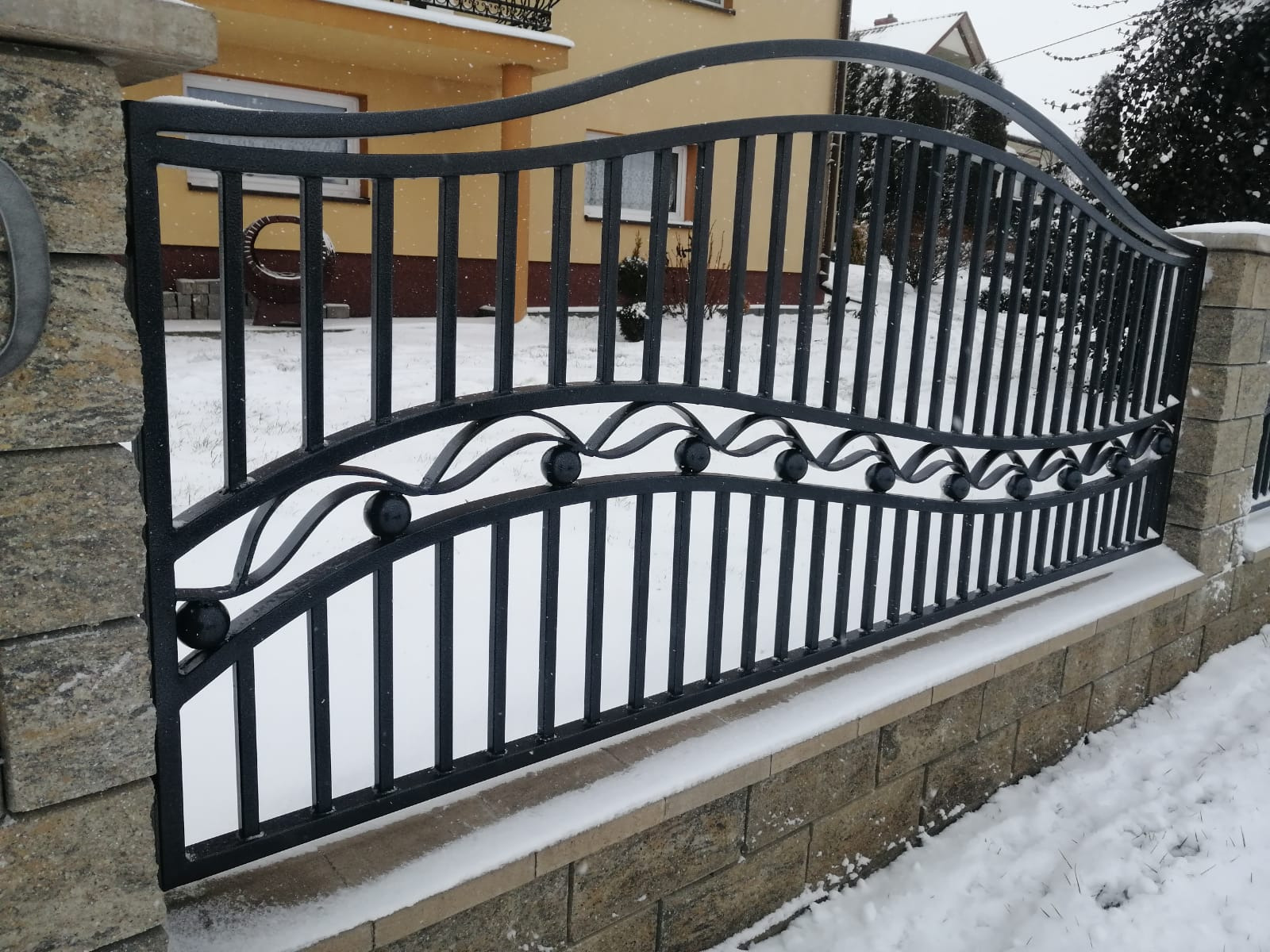 Czarna, metalowa brama z dekoracyjnymi elementami w kształcie kulek, zamontowana na kamiennych słupkach i podmurówce, pokryta śniegiem, na tle ośnieżonego ogrodu i domu z żółtą elewacją.