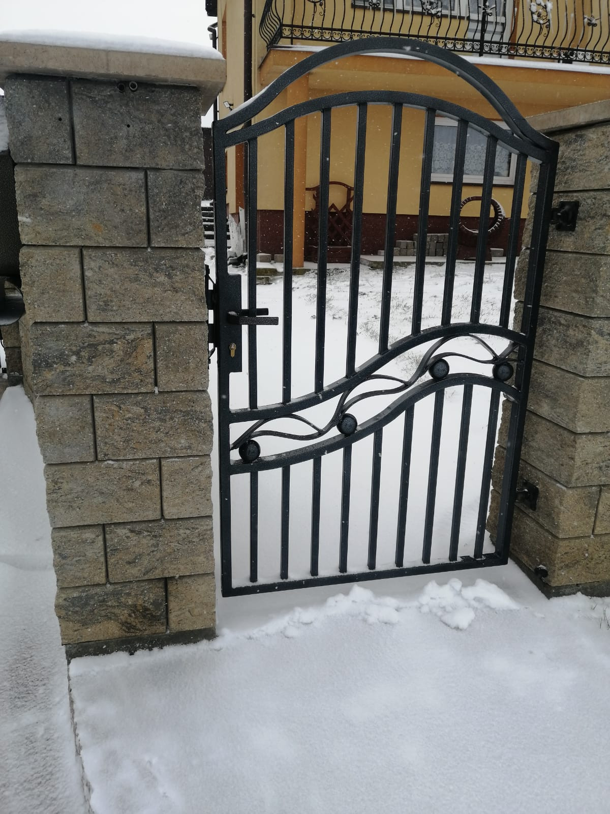 Otwarta, czarna, metalowa furtka wejściowa z dekoracyjnymi elementami, osadzona pomiędzy dwoma kamiennymi słupkami, pokryta śniegiem w zimowej scenerii.