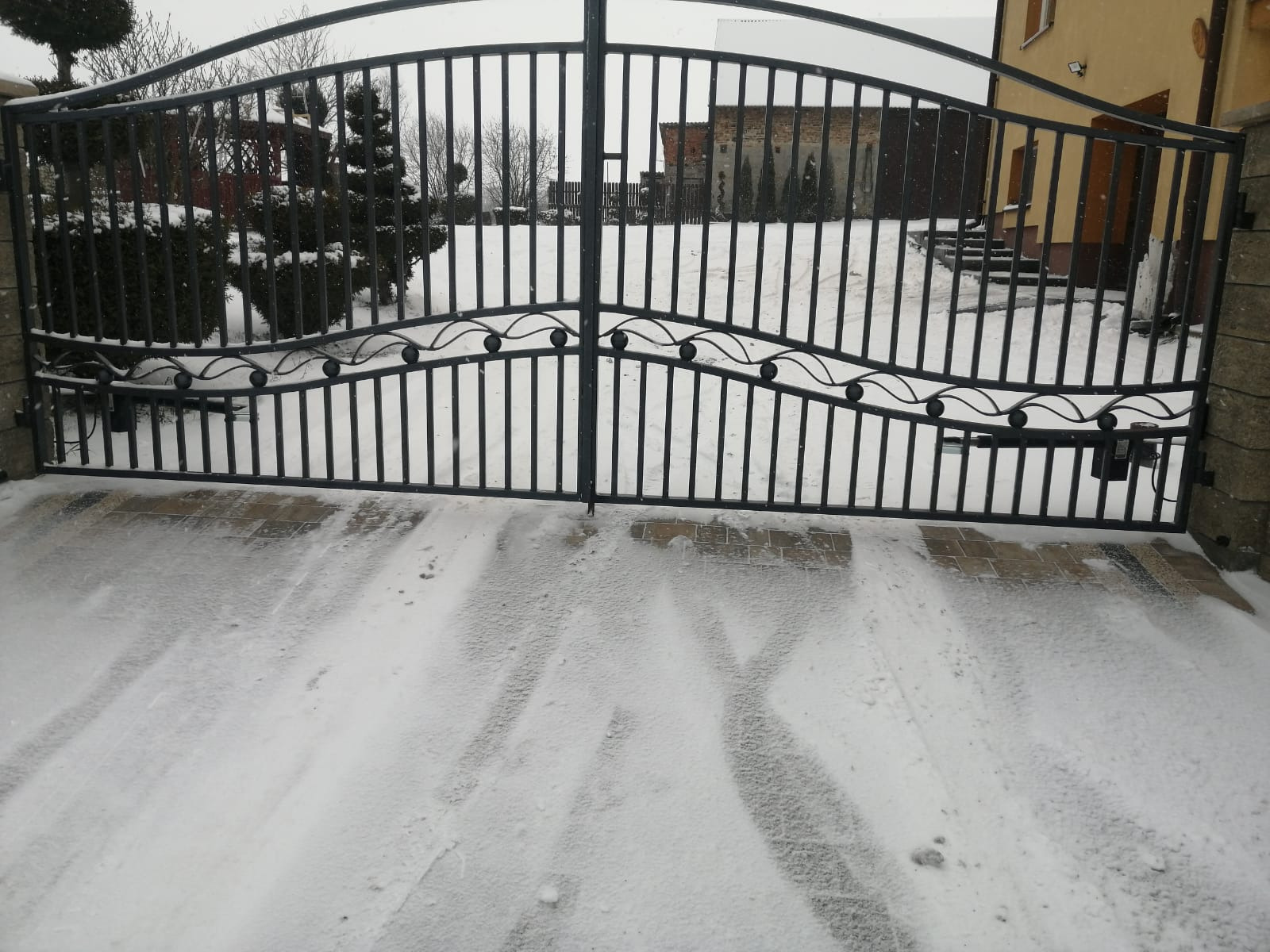Czarna metalowa brama wjazdowa z dekoracyjnymi elementami w kształcie kulek, widok z zewnątrz na ośnieżony podjazd, w tle budynek z czerwonej cegły i zielone drzewa.