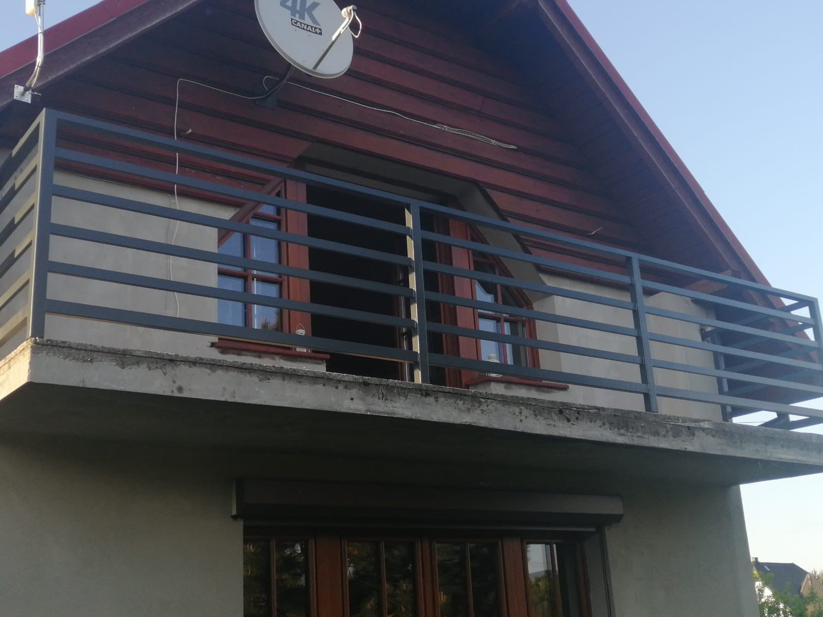 Szara, metalowa balustrada balkonowa z poziomymi szczeblami, zamontowana na betonowej płycie balkonu domu z czerwonym dachem i anteną satelitarną.