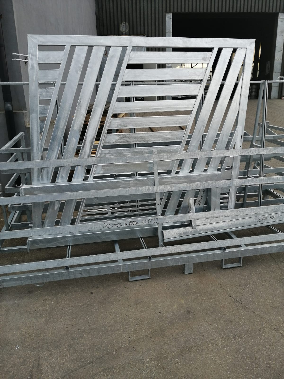 Stalowa, surowa konstrukcja bramy wjazdowej z ukośnymi i poziomymi poprzeczkami, częściowo złożona na palecie transportowej z napisem 'LENOX' wykonanym markerem.