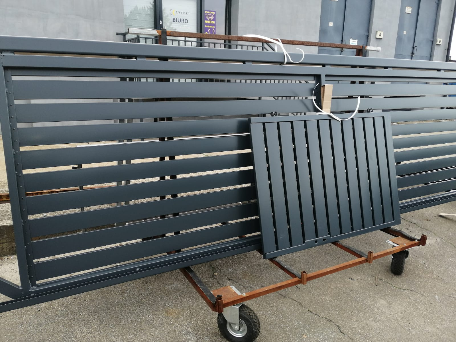 Fragment przesuwnej, metalowej bramy wjazdowej w kolorze grafitowym, z poziomymi panelami, umieszczony na wózku transportowym, w tle budynek z szarą elewacją.