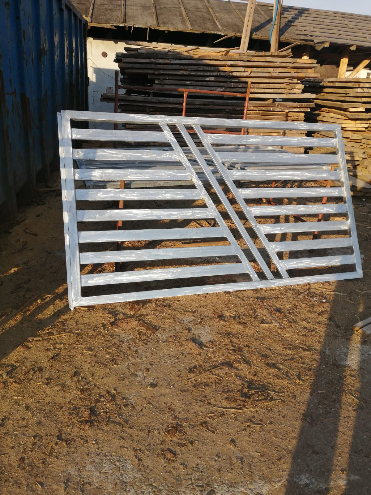 Surowa, metalowa rama bramy z poziomymi szczeblami i ukośnymi wzmocnieniami, leżąca na ziemi przed stertą desek i niebieskim kontenerem.
