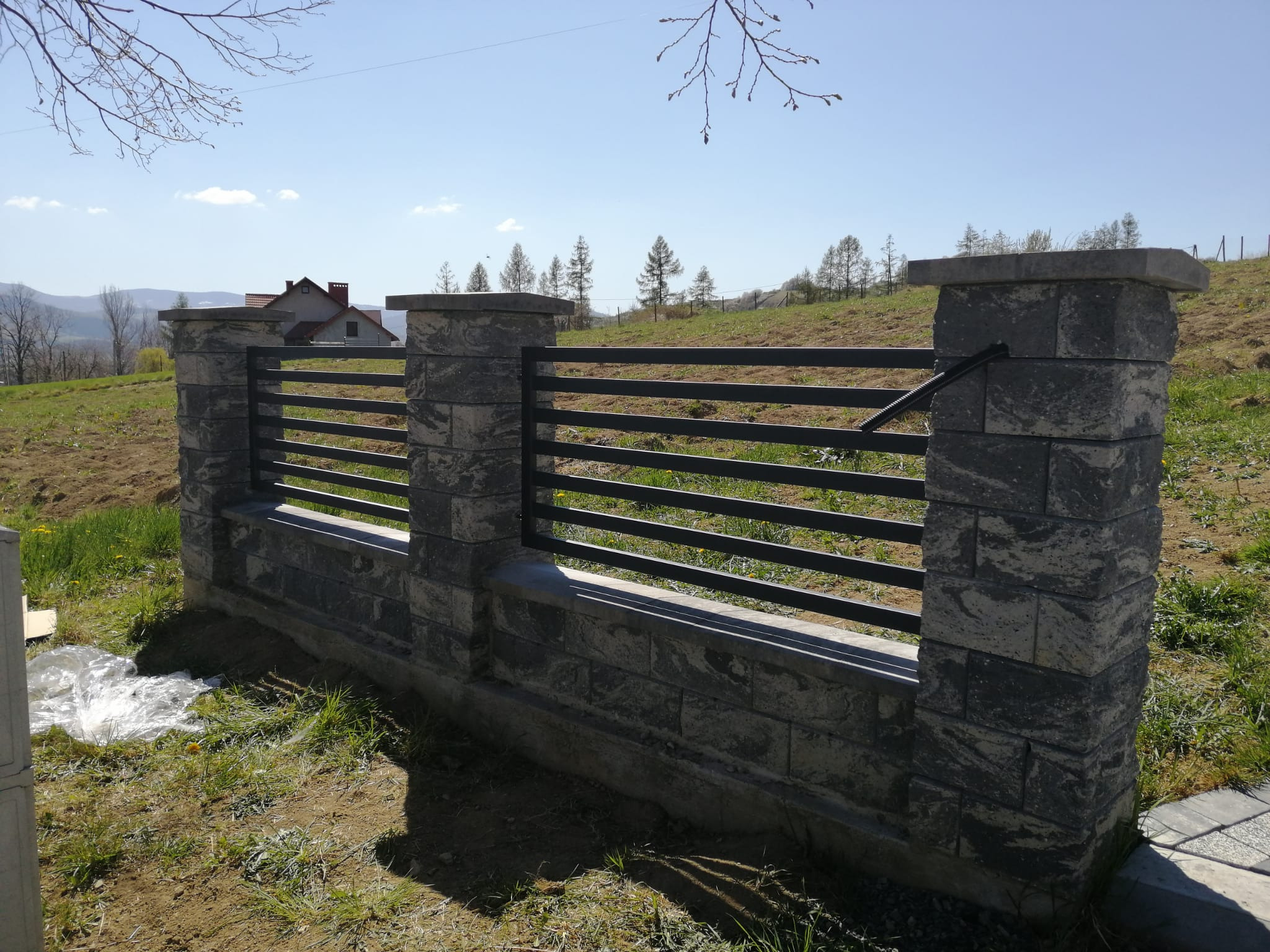 Fragment ogrodzenia z metalowymi przęsłami i kamiennymi słupkami na tle pagórkowatego krajobrazu i błękitnego nieba, widoczne ślady prac ziemnych.