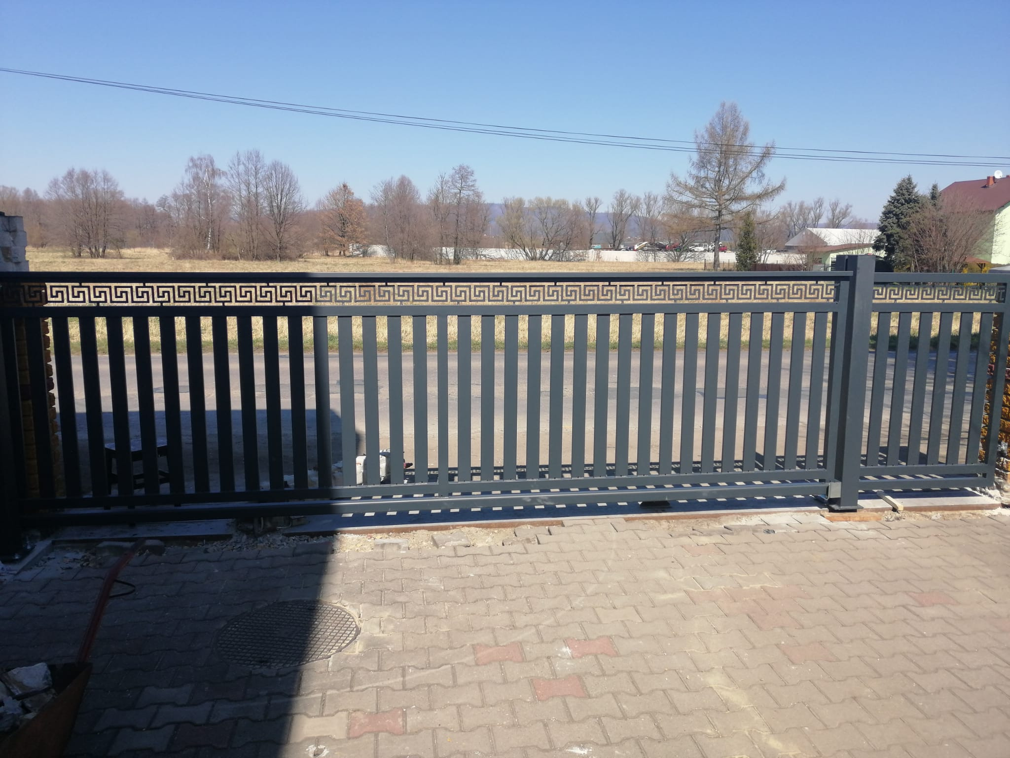 Szara, przesuwna brama wjazdowa z dekoracyjnym, geometrycznym wzorem u góry, zamontowana na posesji z kostką brukową, na tle krajobrazu z drzewami i zabudowaniami.