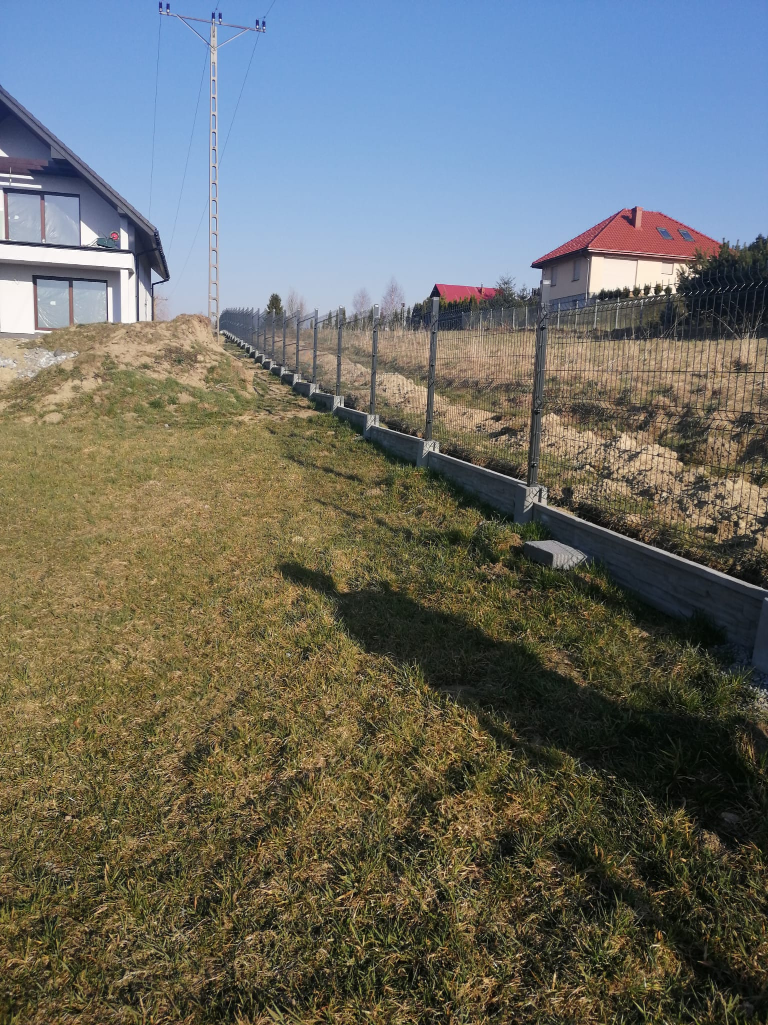 Nowoczesne ogrodzenie panelowe z podmurówką betonową na nierównym terenie, widok z dołu, w tle domy jednorodzinne i słup energetyczny.