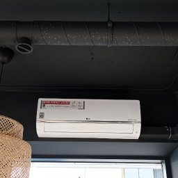 ECOKLIMA SPÓŁKA Z OGRANICZONĄ ODPOWIEDZIALNOŚCIĄ - Biały klimatyzator LG Dual Inverter zamontowany na czarnej ścianie, widoczne przewody wentylacyjne i dekoracyjna lampa z rattanu.