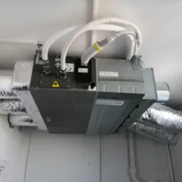 Centrala wentylacyjna z rekuperacją marki Haier zamontowana pod sufitem z widocznymi przewodami wentylacyjnymi w izolacji aluminiowej i białych rurach doprowadzających powietrze.