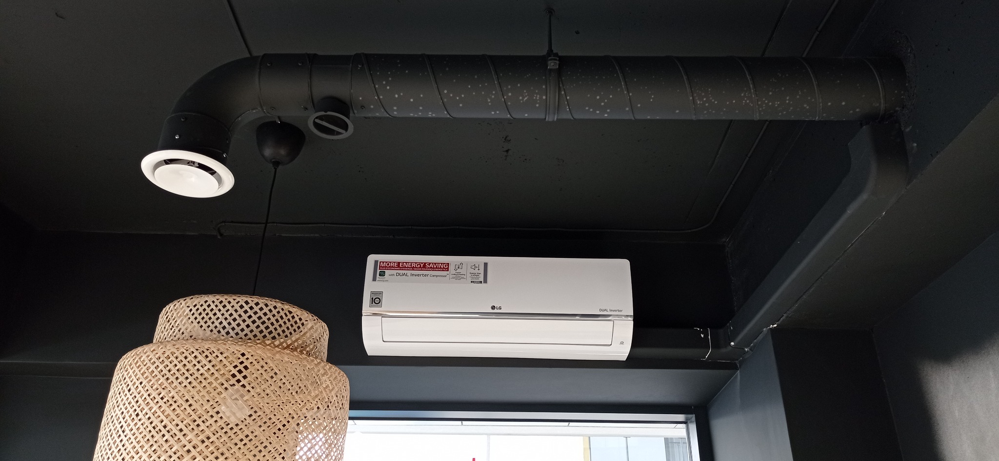Biały klimatyzator LG Dual Inverter zamontowany na czarnej ścianie, widoczne przewody wentylacyjne i dekoracyjna lampa z rattanu.
