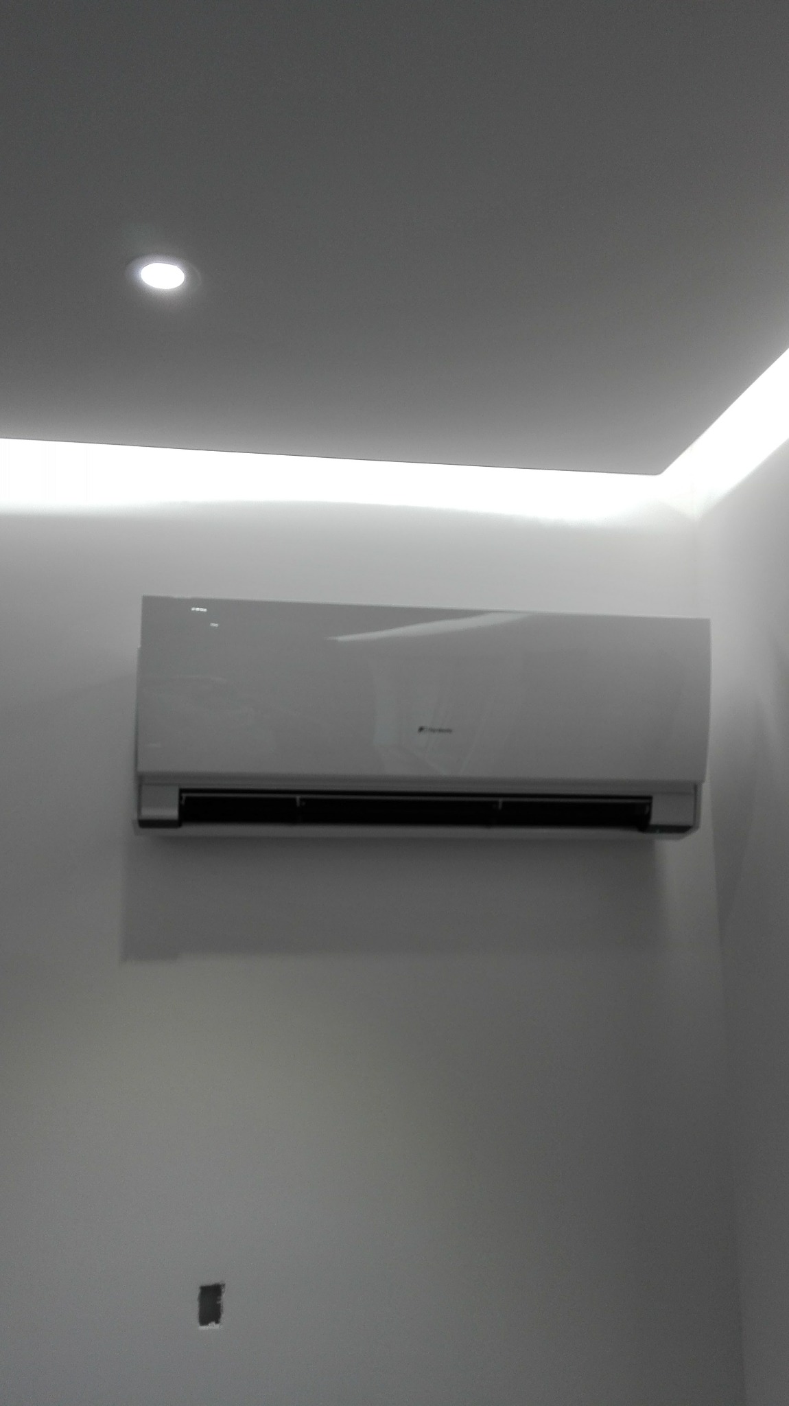 Biała jednostka klimatyzacyjna zamontowana na ścianie w pomieszczeniu z oświetleniem LED na suficie i punktowym światłem. Widoczny otwór na gniazdko elektryczne poniżej.