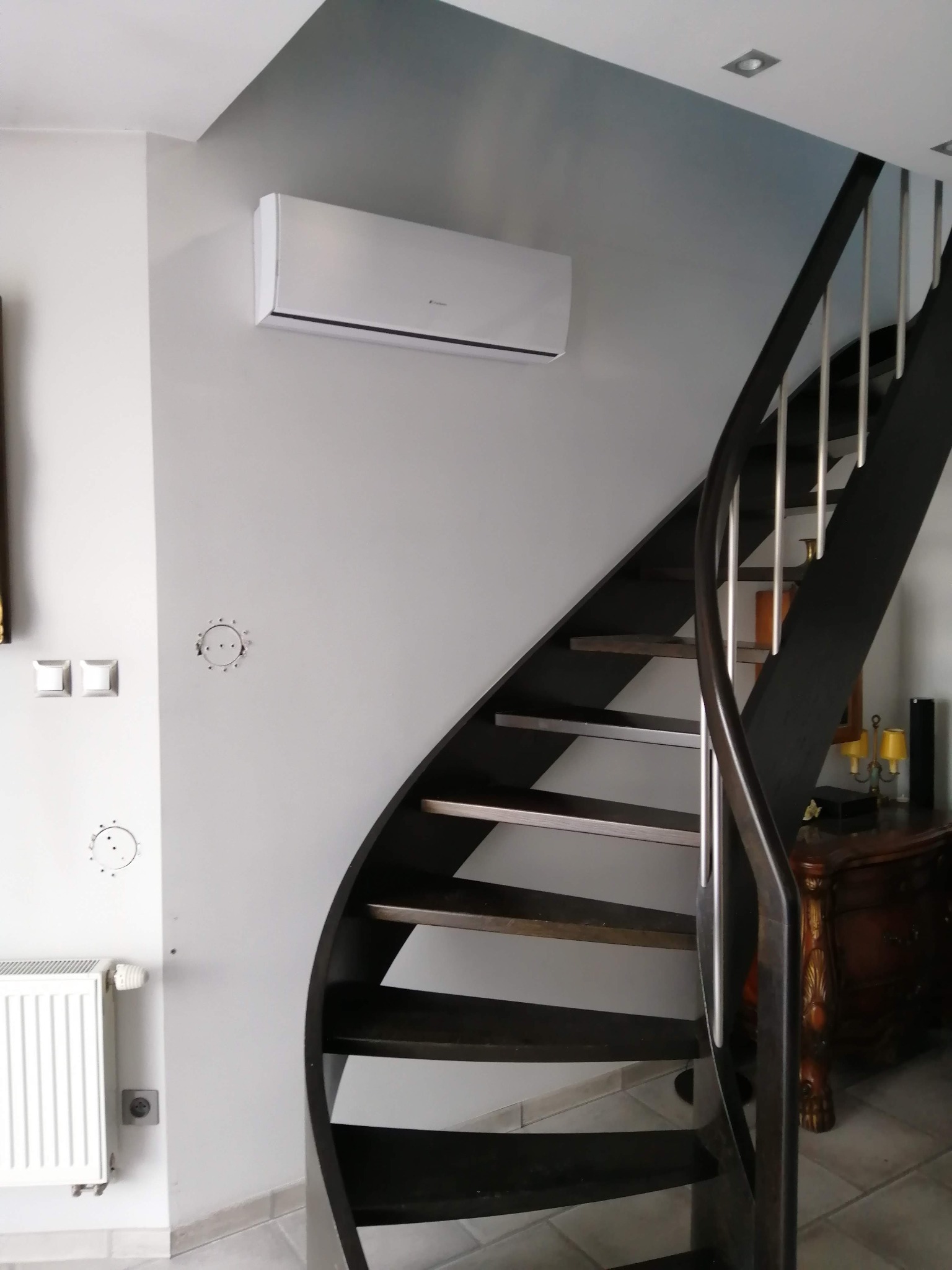 Biały klimatyzator ścienny zamontowany w domu obok kręconych schodów, widoczne ślady po poprzednich instalacjach na ścianie.