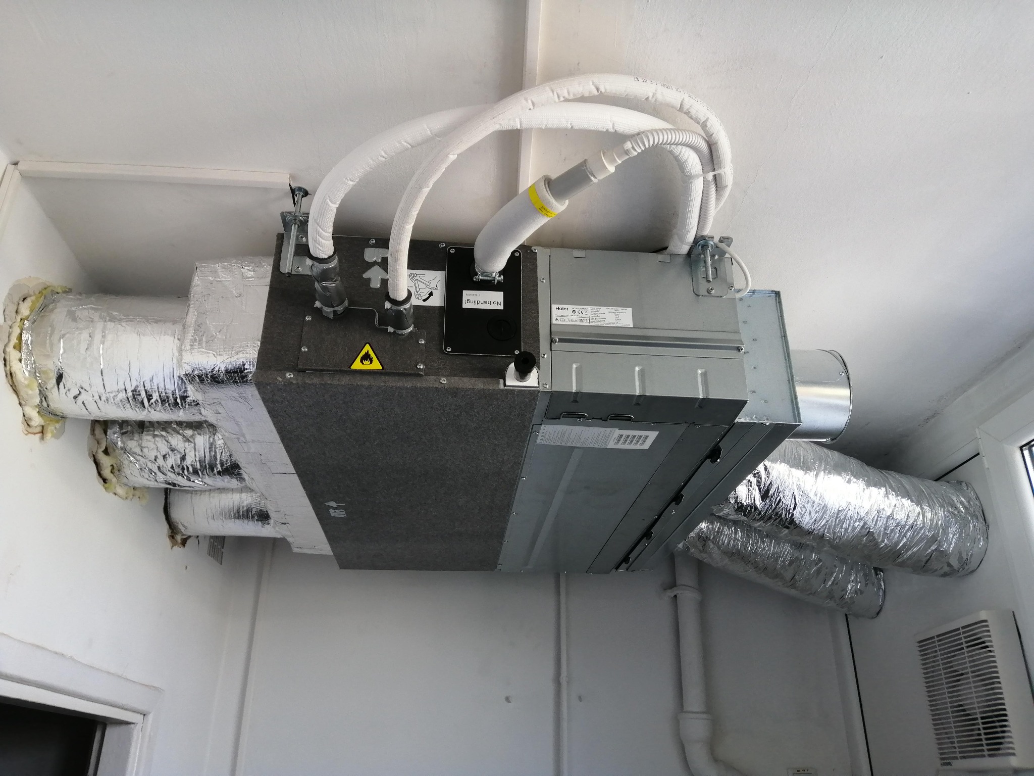 Centrala wentylacyjna z rekuperacją marki Haier zamontowana pod sufitem z widocznymi przewodami wentylacyjnymi w izolacji aluminiowej i białych rurach doprowadzających powietrze.