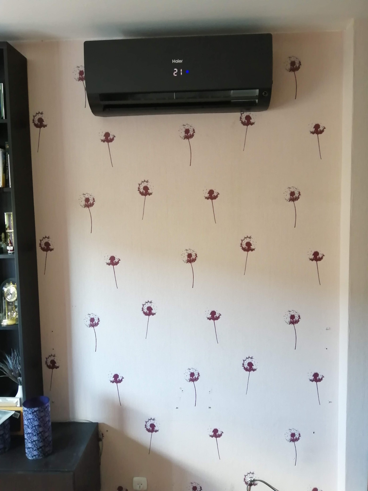 Czarny klimatyzator Haier zamontowany na ścianie z motywem dmuchawców, wyświetlający temperaturę 21 stopni Celsjusza.