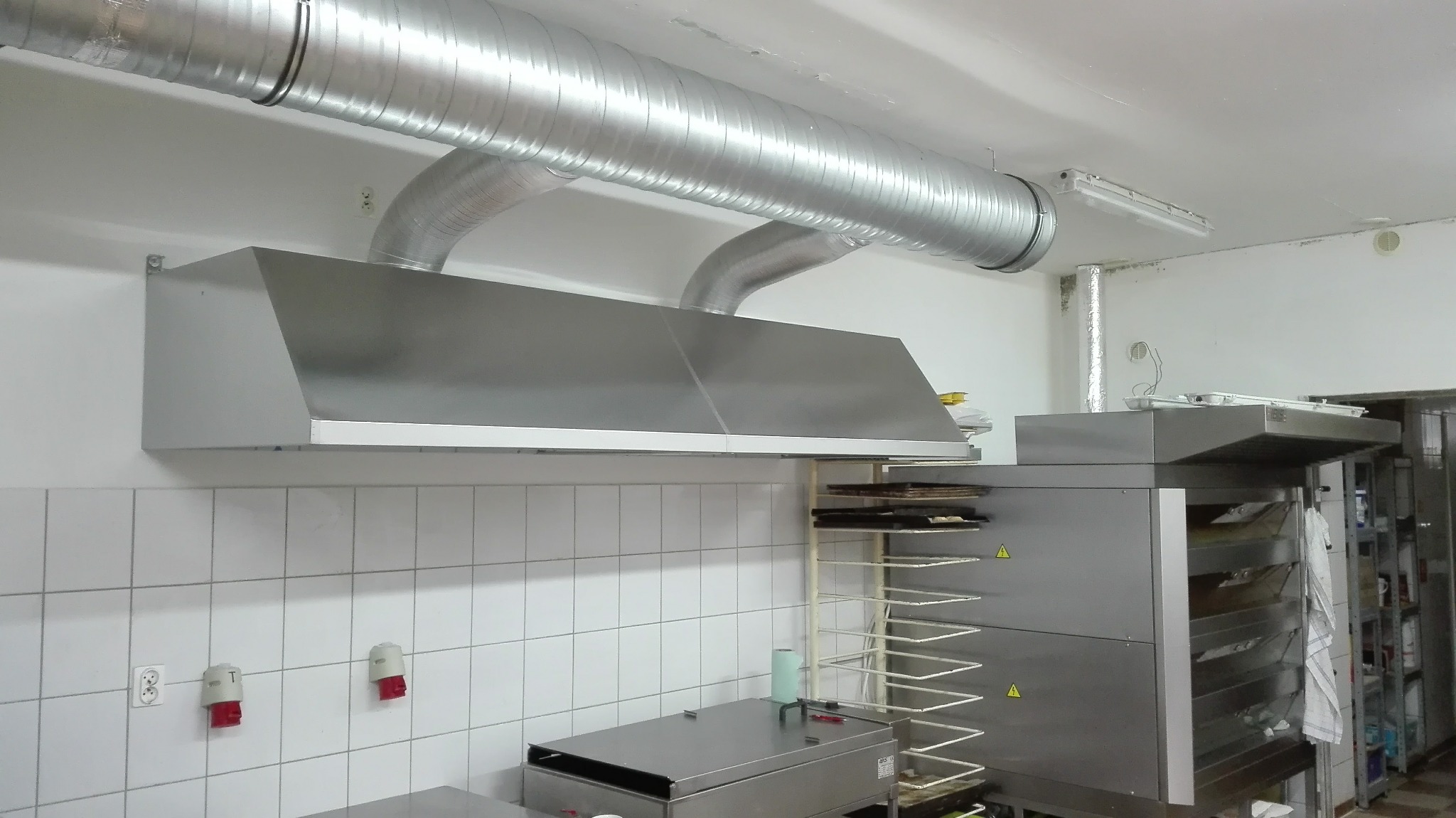 Kuchenny okap przemysłowy ze stali nierdzewnej z podwieszonym systemem wentylacyjnym, widoczne gniazda elektryczne i włączniki na kafelkowej ścianie.