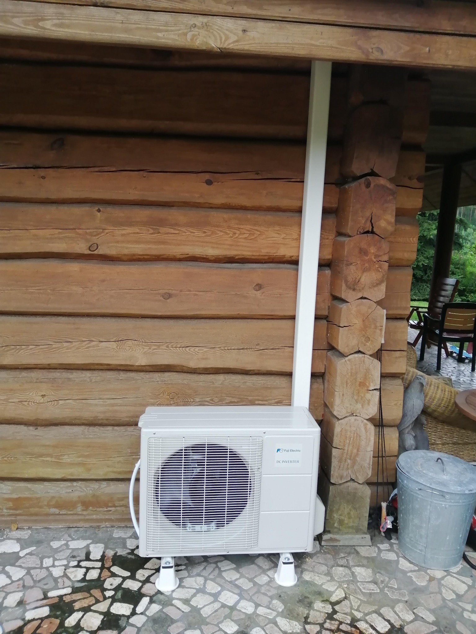 Zewnętrzna jednostka klimatyzacji Fuji Electric DC Inverter, zamontowana na białych wspornikach przy drewnianej ścianie z bali, widoczna rura odprowadzająca oraz metalowy kosz na śmieci w tle.