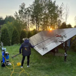 Czyszczenie paneli słonecznych przez osobę z użyciem specjalistycznego sprzętu na tle zieleni i zachodzącego słońca. Praca wykonywana z ziemi.