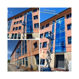 PREMO AGNIESZKA KONOPCZYK - Pracownik na podnośniku koszowym myje okna budynku, odbijające błękitne niebo. Używana woda demineralizowana, widoczne urządzenie do mycia i kciuk w górę.