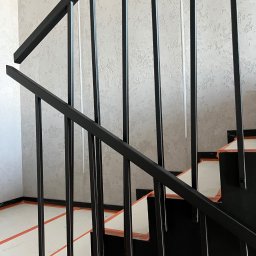 PREMO AGNIESZKA KONOPCZYK - Czarne metalowe schody z zabezpieczeniem folią i taśmą, widok z dołu, ściana z teksturą w tle. Minimalistyczny design, zabezpieczenie przed zabrudzeniem podczas prac wykończeniowych.