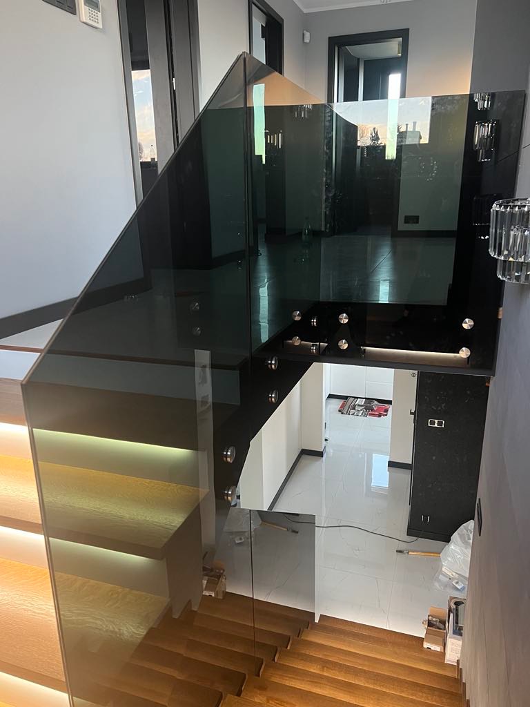 Nowoczesne schody z drewnianymi stopniami i szklaną balustradą, oświetlone od spodu, widok z góry na jasne wnętrze domu.