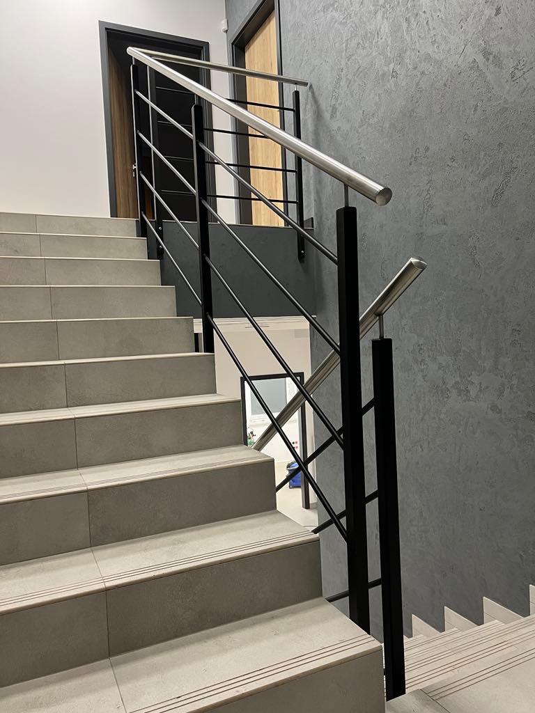Schody z szarych płytek z czarną metalową balustradą i srebrną poręczą. Widok od dołu, drzwi w tle.