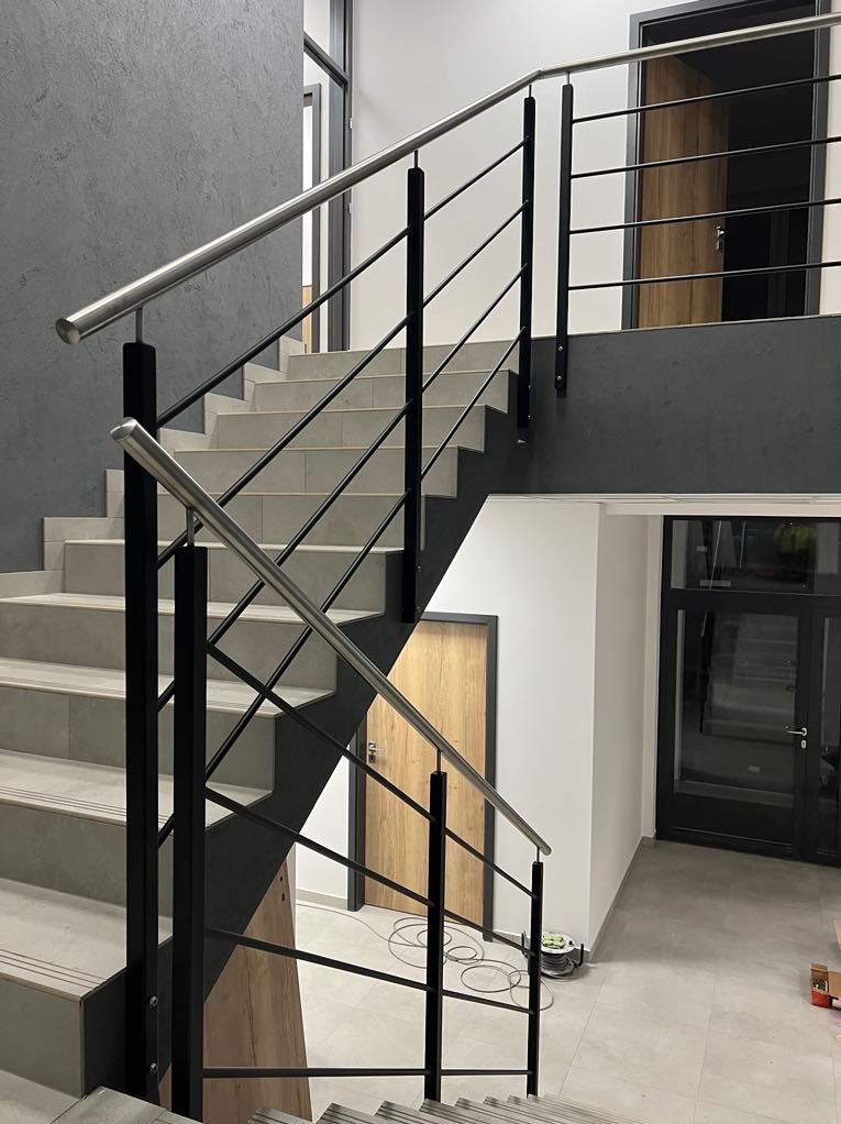 Nowoczesne schody z metalową balustradą w minimalistycznym wnętrzu. Szare płytki schodowe i czarne elementy balustrady kontrastują z jasnymi ścianami i drewnianymi drzwiami.