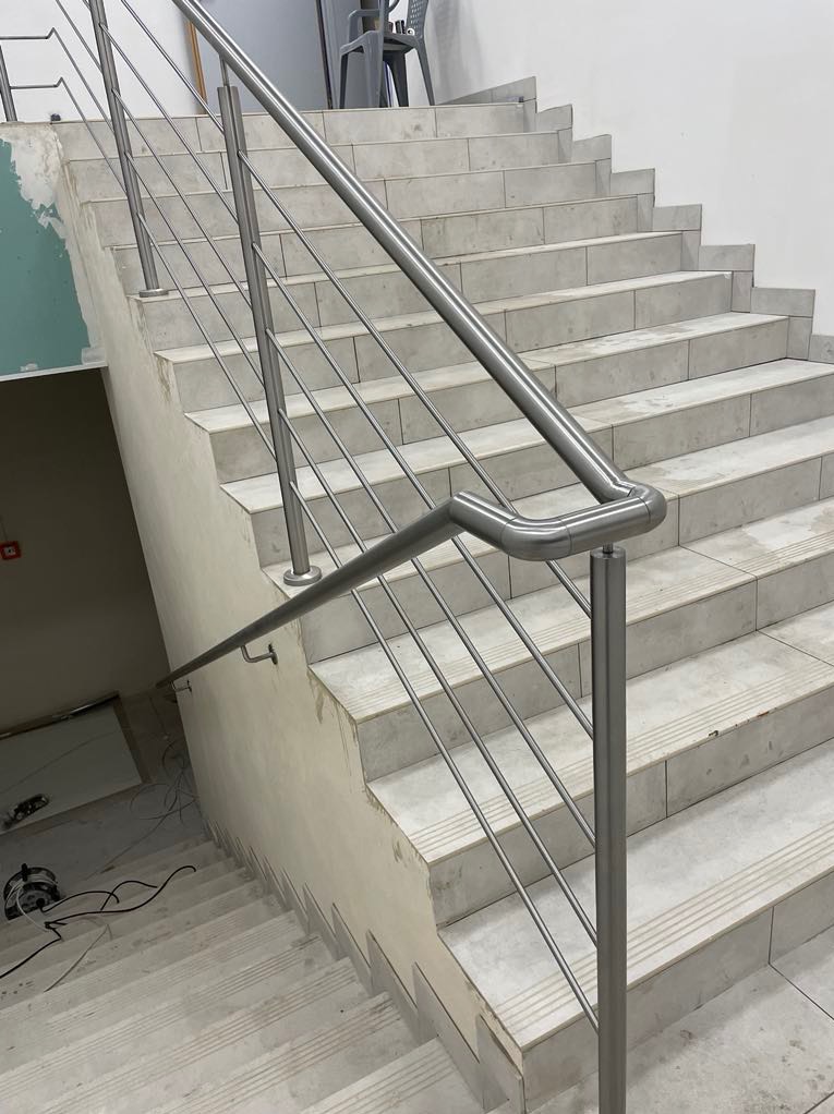 Schody wyłożone płytkami z zamontowaną, nowoczesną balustradą ze stali nierdzewnej. Widoczne niedokończone prace wykończeniowe.