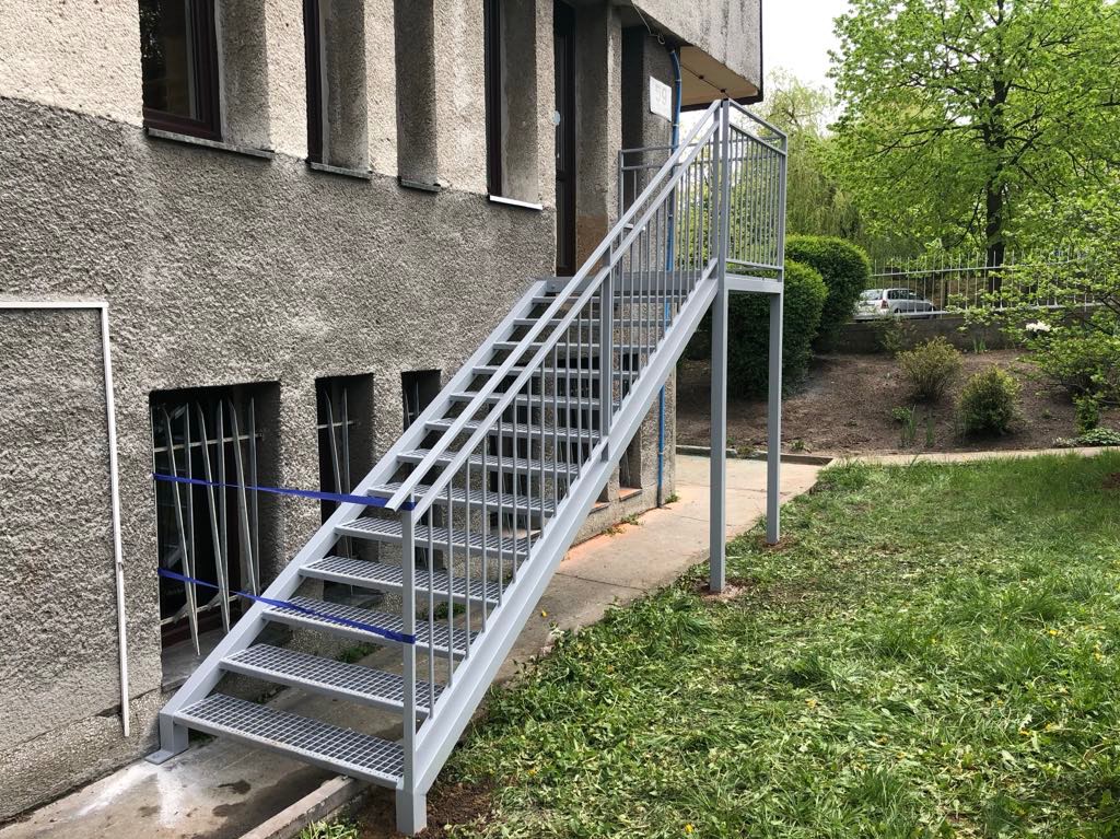 Zewnętrzne, metalowe schody z ażurowymi stopniami, prowadzące do budynku z szarą elewacją. Widoczne trawnik i zieleń w tle. Ujęcie z dołu, podkreślające konstrukcję.