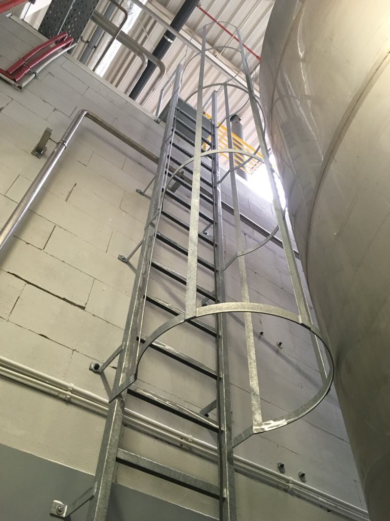 Metalowa drabina przymocowana do ściany z bloków betonowych, prowadząca do zbiornika. Widoczne rury instalacyjne. Ujęcie od dołu, podkreślające wysokość konstrukcji.