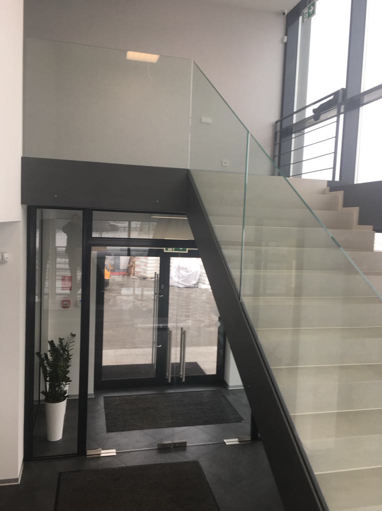 Wnętrze budynku z nowoczesną klatką schodową z szklaną balustradą i przeszklonym wejściem. Widoczna roślina doniczkowa i wycieraczka przy wejściu. Stonowana kolorystyka.