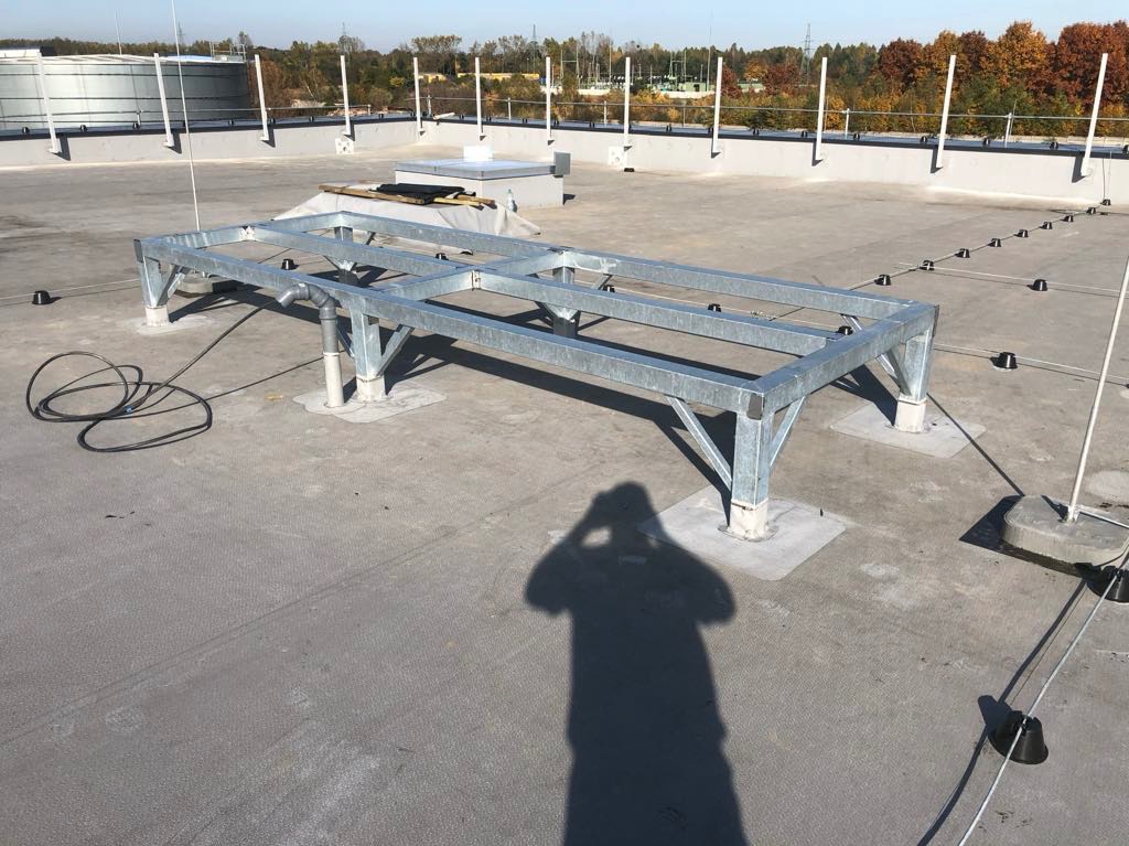 Metalowa konstrukcja wsporcza na płaskim dachu z widocznym systemem odgromowym i shadow osoby robiącej zdjęcie. Widok z góry, w tle drzewa i zbiornik.