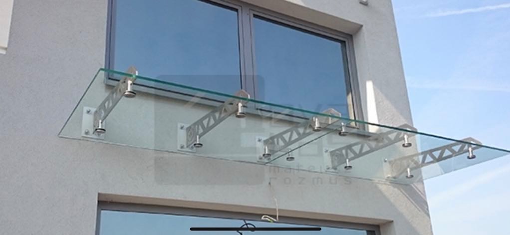 Szklany daszek nad wejściem, wsparty na metalowych wspornikach, zamocowany do elewacji budynku. Widoczne okno i fragment drugiego okna poniżej.