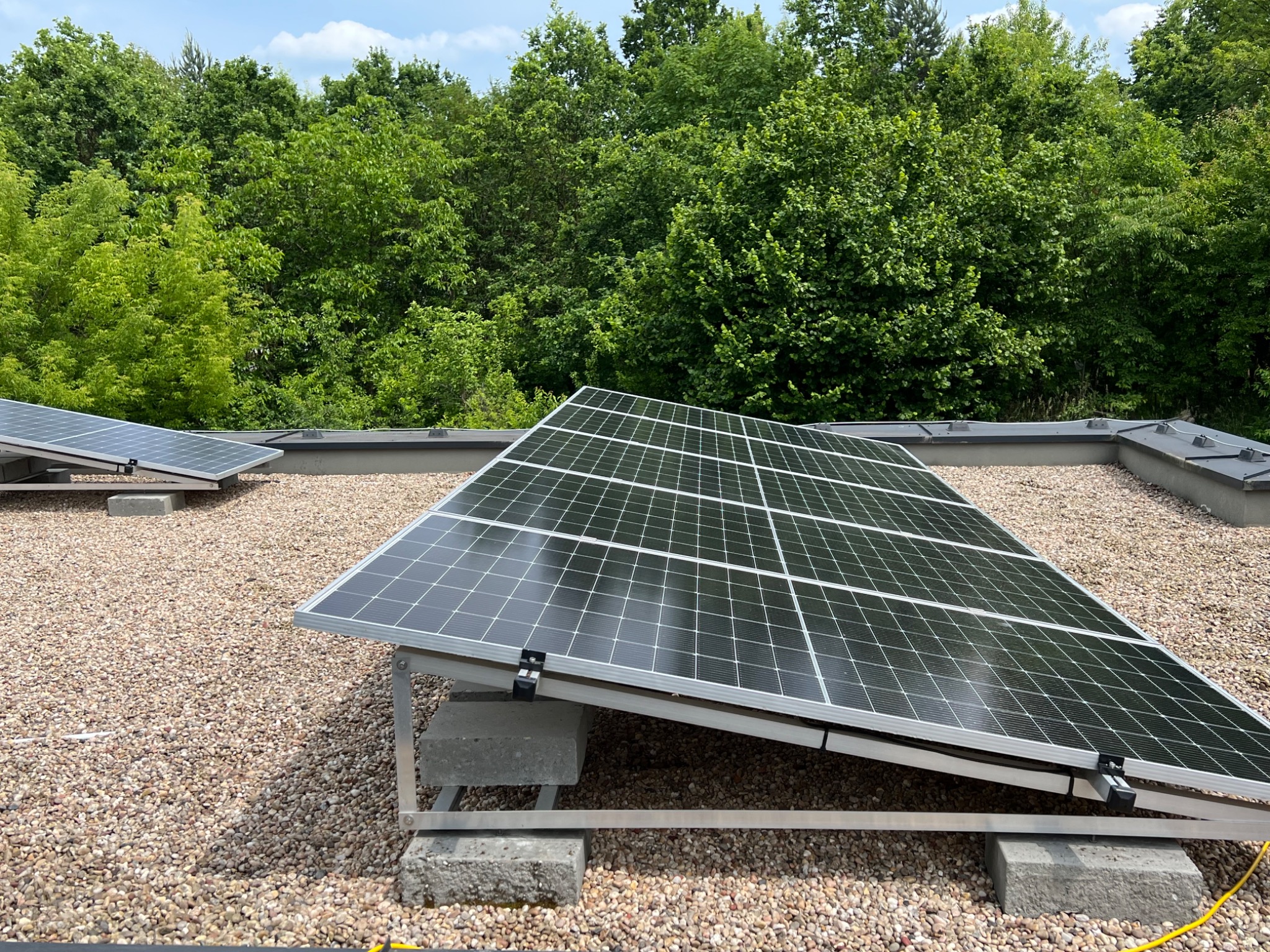 Panel słoneczny na dachu z gruzem, wsparty na betonowych blokach, otoczony zielenią drzew. Perspektywa z dołu ukazuje instalację i tło.