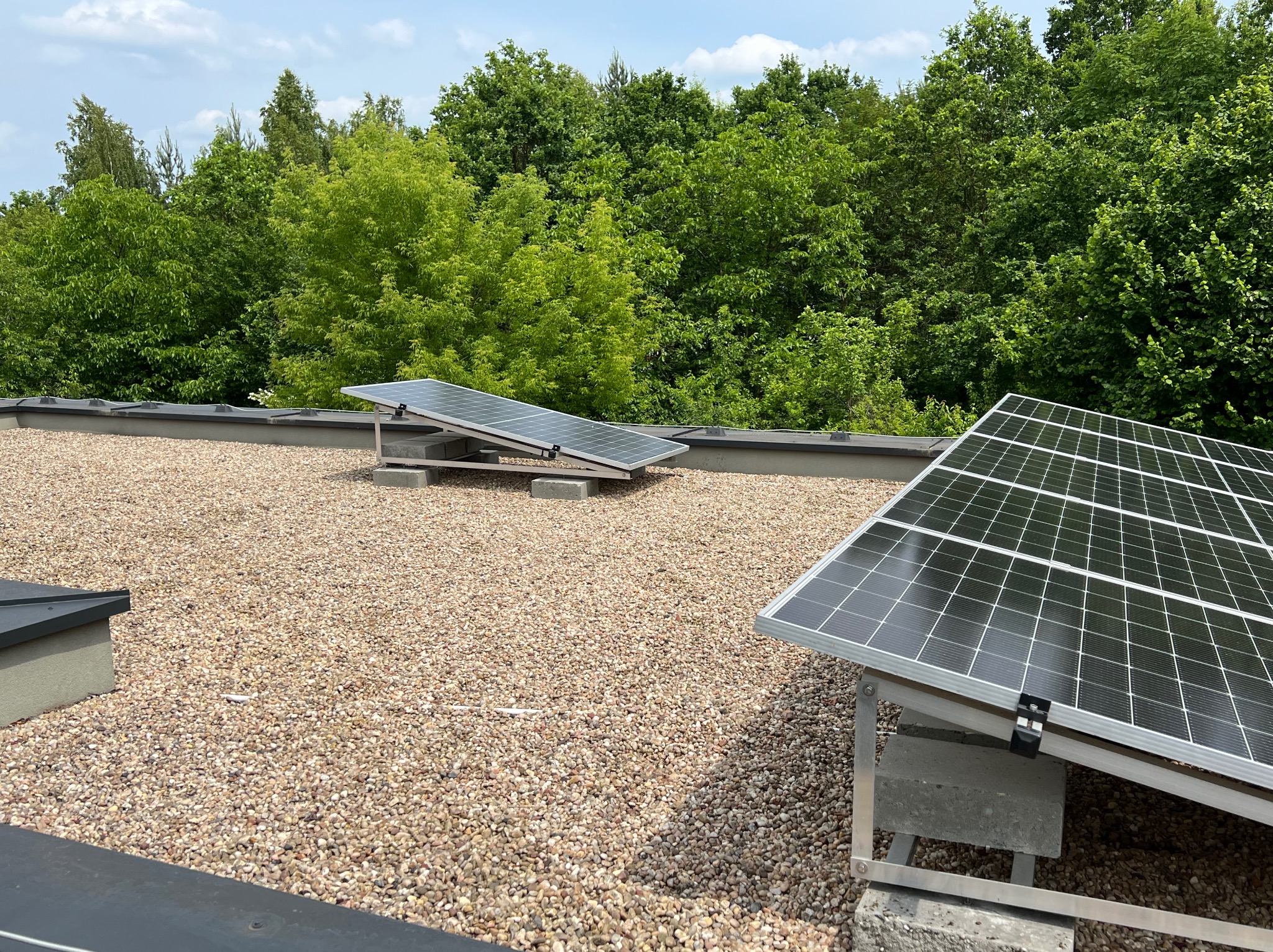 Panele słoneczne na płaskim dachu pokrytym drobnym żwirem, z zielenią drzew w tle. Metalowe konstrukcje wsporcze paneli, betonowe podstawy. Widok z góry, słoneczny dzień.