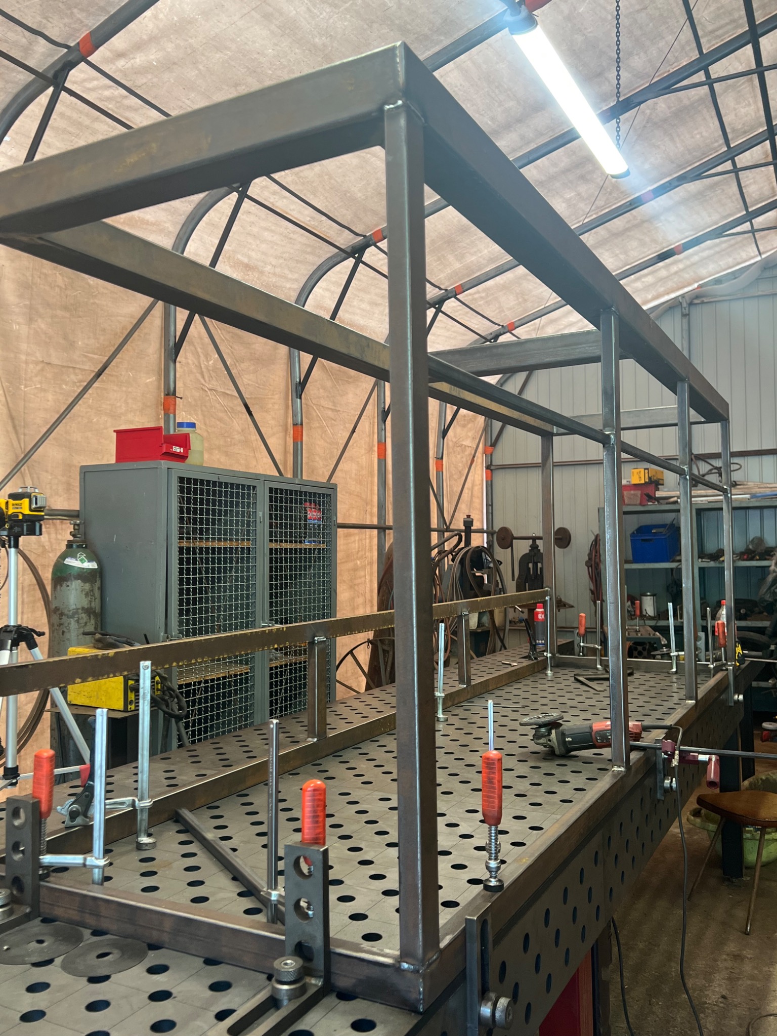 Metalowa konstrukcja szkieletowa w warsztacie, elementy mocowane zaciskami. Widoczny stół spawalniczy z otworami, narzędzia i szafka warsztatowa w tle.