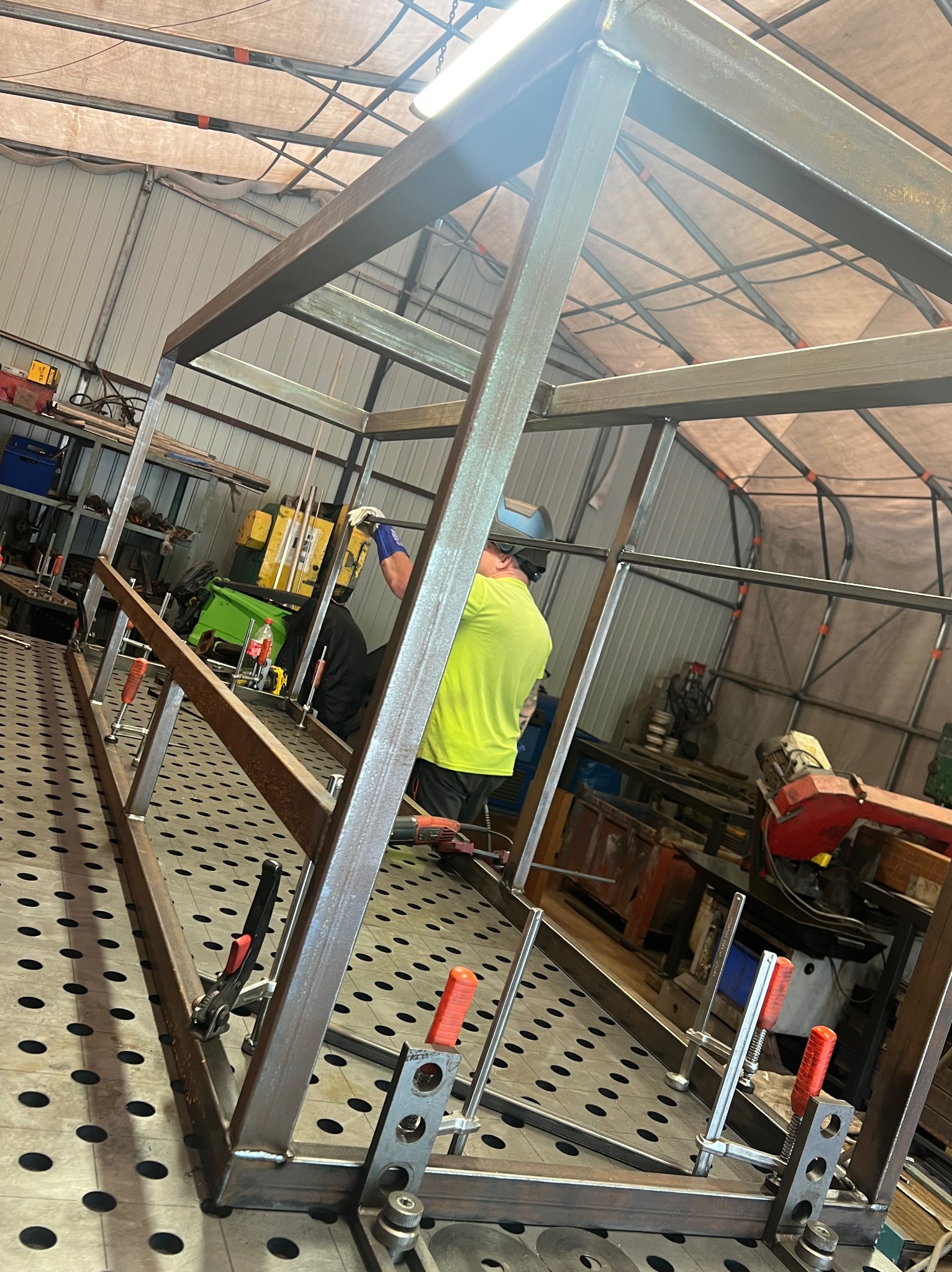 Spawacz łączy metalowe profile w ramę na perforowanym stole spawalniczym, używając zacisków. Widoczne narzędzia i osłona spawalnicza.