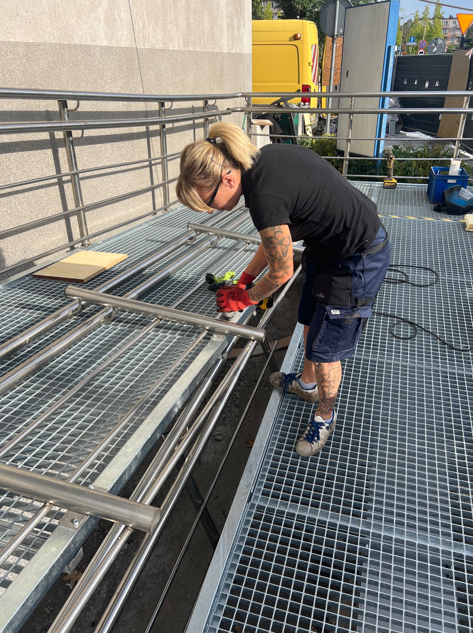 Pracownik w Konopiskach szlifuje metalową konstrukcję ogrodzenia. Widoczne iskry, rękawice ochronne i elementy metalowe. Praca na zewnątrz.