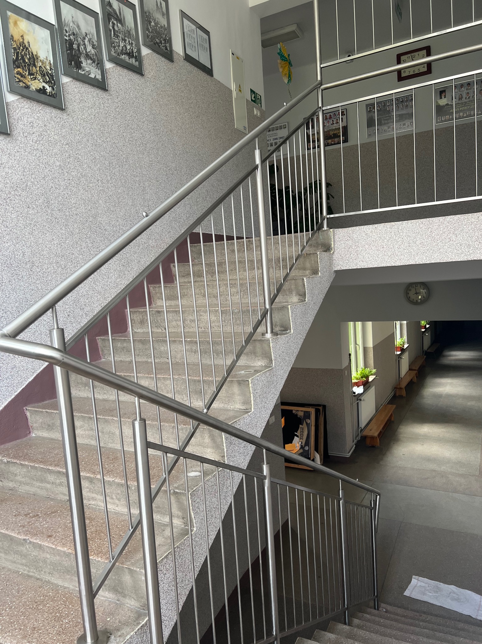 Schody z metalową balustradą w budynku użyteczności publicznej, z obrazami na ścianach i widokiem na korytarz z ławkami i roślinami w donicach.