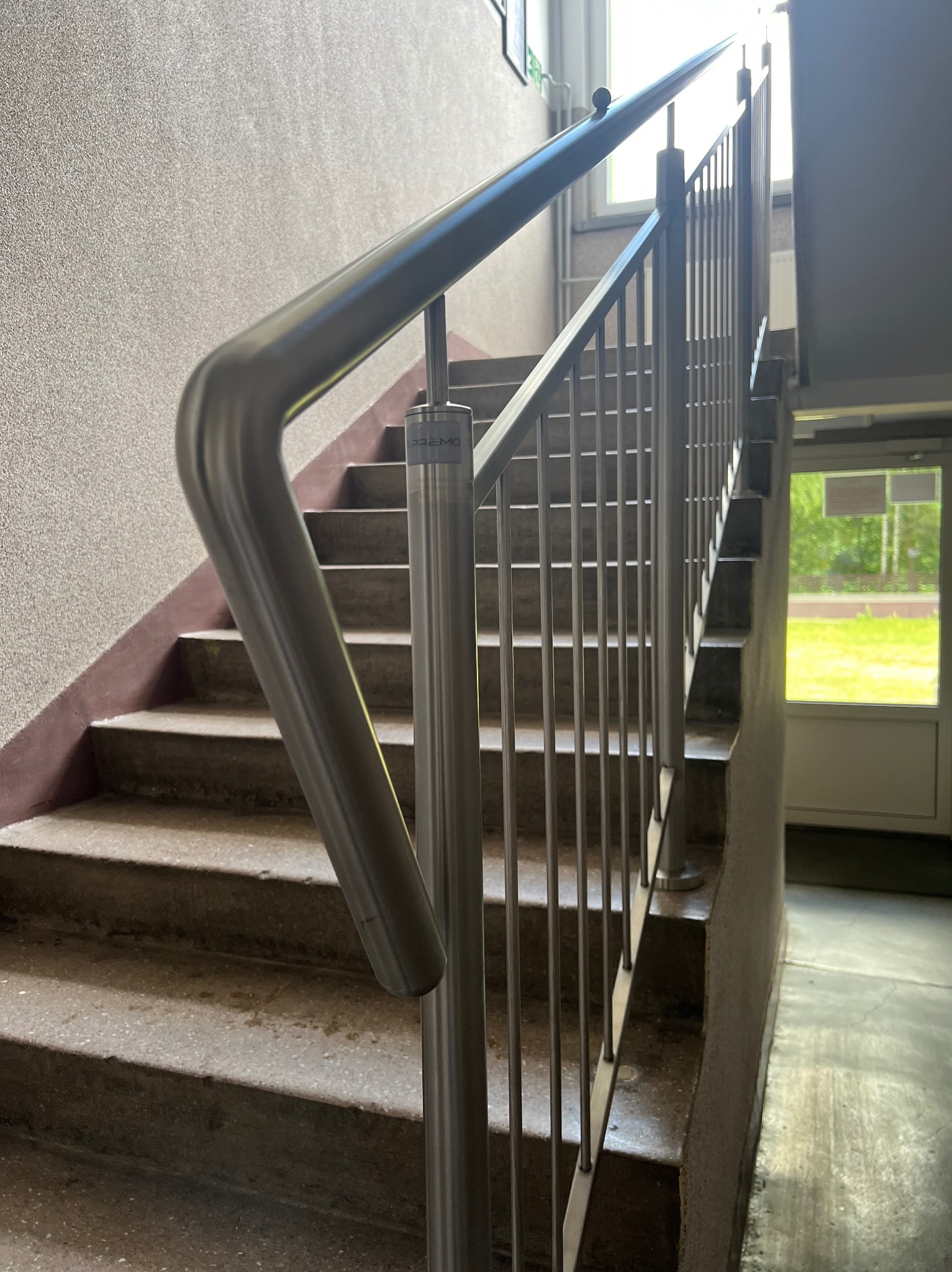 Schody betonowe z metalową, nowoczesną balustradą. Widok z dołu, w tle okno z zielenią. Poręcz ze stali nierdzewnej z zaokrąglonym zakończeniem.