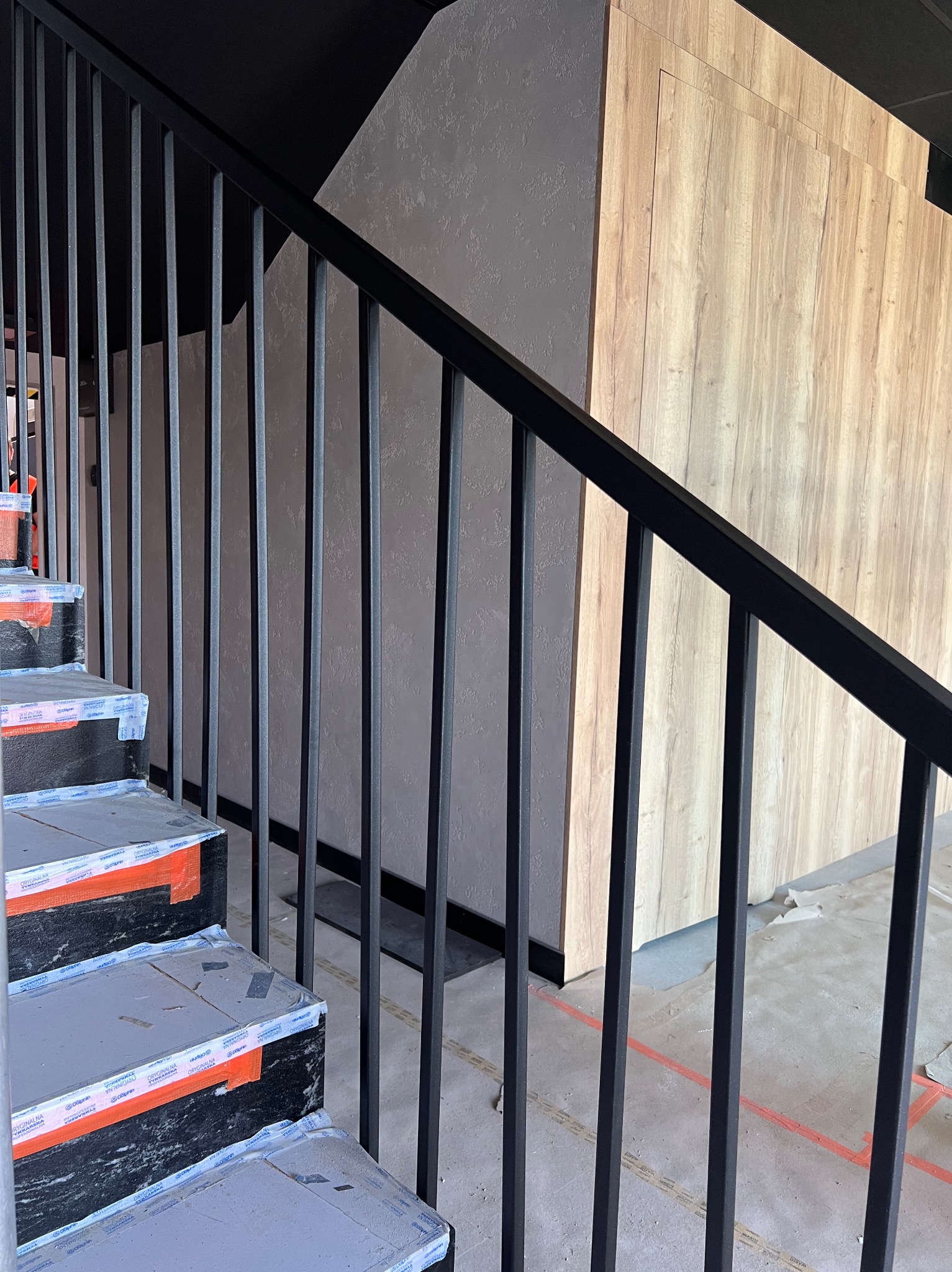 Nowoczesne, czarne metalowe schody z zabezpieczonymi stopniami w trakcie montażu, na tle ściany z elementami drewnopodobnymi.