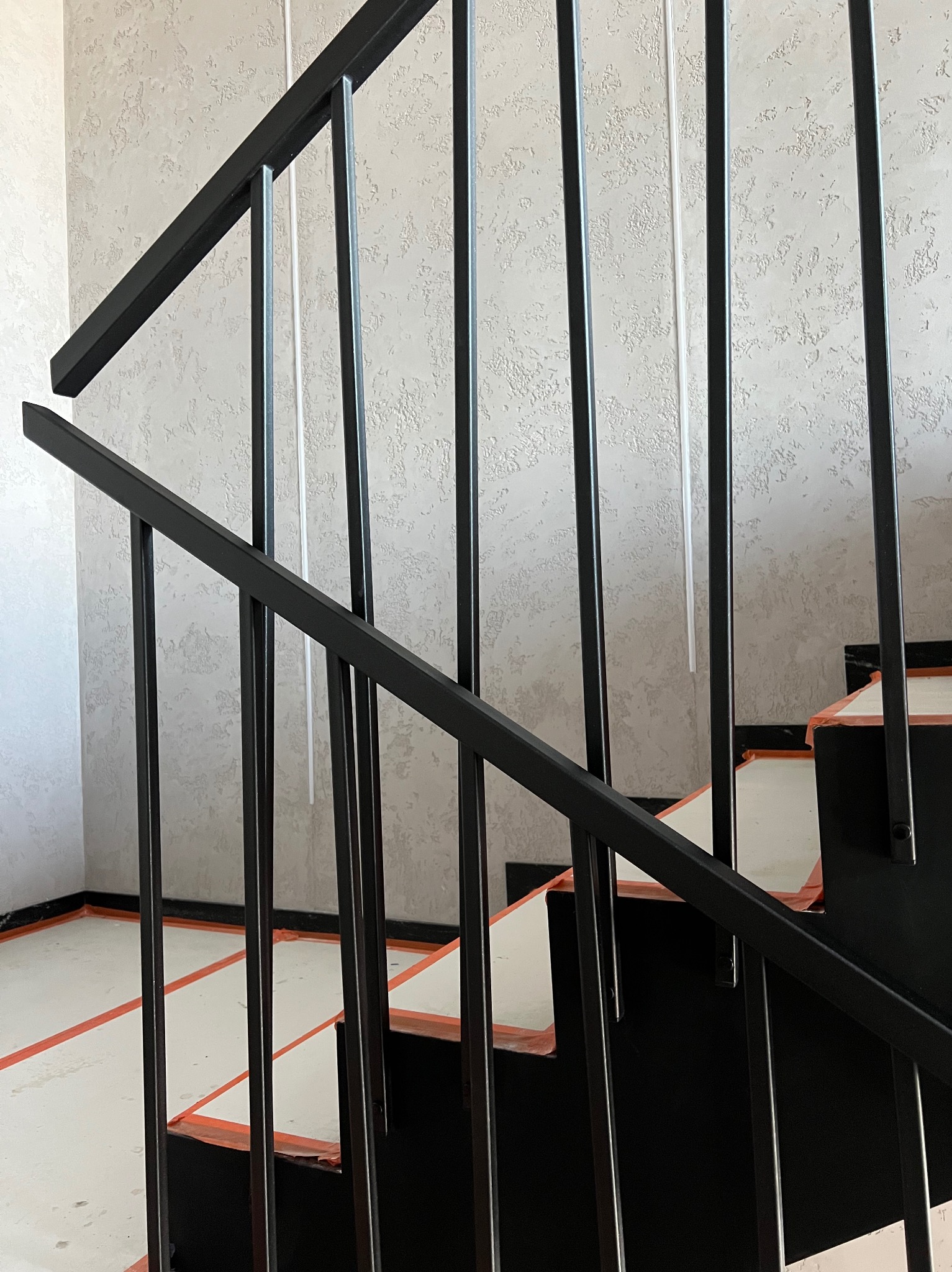 Czarne metalowe schody z zabezpieczeniem folią i taśmą, widok z dołu, ściana z teksturą w tle. Minimalistyczny design, zabezpieczenie przed zabrudzeniem podczas prac wykończeniowych.