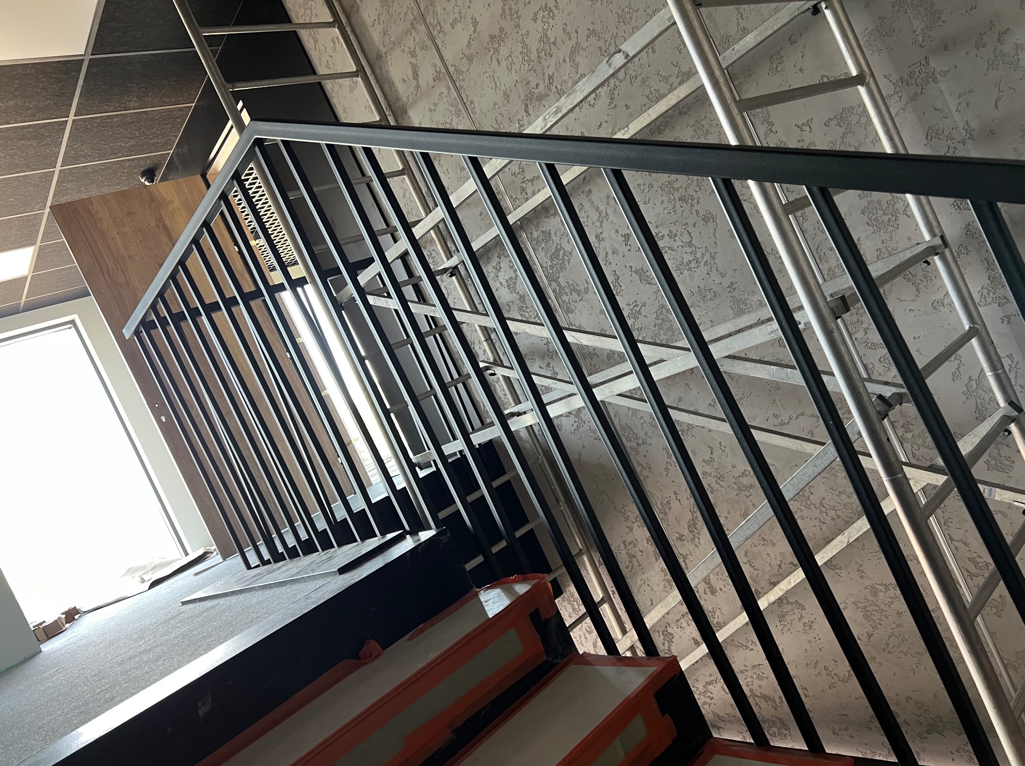 Nowoczesna, czarna balustrada schodowa z metalowymi prętami, widoczna z dołu pod kątem, z fragmentem rusztowania w tle i oknem wpadającym światłem.